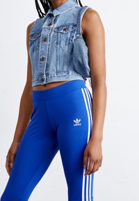 Chaleco de mezclilla recortado con cuello y cierre de botones, combinado con leggins azules que tienen rayas blancas de Adidas y el logo en la cadera.