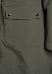 Olive zelena tkanina z velikim pravokotnim žepom, ki je pritrjen z dvema črnima gumboma.