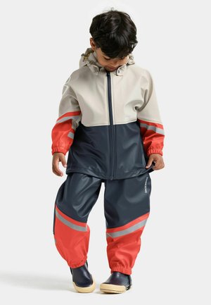 Ensemble veste et pantalon imperméables en matériau synthétique. Les couleurs comprennent beige, bleu marine et rouge vif avec des bandes réfléchissantes.