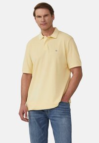 Koszulka polo w miękkim żółtym bawełnianym materiale z klasycznym kołnierzykiem, krótkimi rękawami i małym haftowanym logo na piersi; zestawiona z niebieskimi dżinsami.