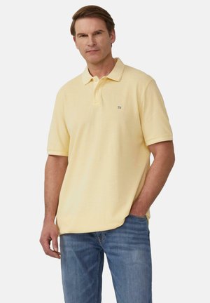 Poloshirt aus weichem gelbem Baumwollstoff mit klassischem Kragen, kurzen Ärmeln und einem kleinen bestickten Logo auf der Brust; kombiniert mit blauen Jeans.