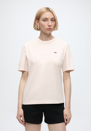 Kobieta o krótkich blond włosach, ubrana w jasnobeżowy t-shirt z małym logo i czarne krótkie spodenki, stojąca na jednolitym białym tle.