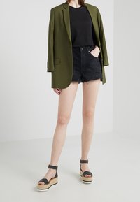 Mujer llevando un blazer verde oliva sobre una camiseta negra y pantalones cortos de mezclilla negra desgastados, junto con sandalias de plataforma con tiras al tobillo en negro.