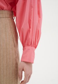Blusa rosa con maniche a palloncino e polsini arricciati, abbinata a una gonna beige con texture a spina di pesce, mostrando un netto contrasto di colore e tessuto.