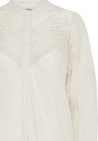 Blusa color crema con chiusura a bottoni, delicato pizzo, tessuto testurizzato con piccoli puntini in rilievo e colletto alla cinese.