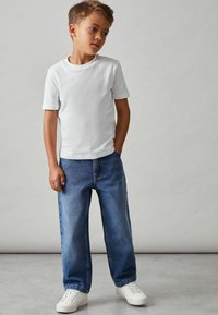 Chlapec v jednoduchém bílém tričku a modrých džínech s volným střihem, doplněný bílými teniskami; ruce v kapsách, neutrální pozadí.