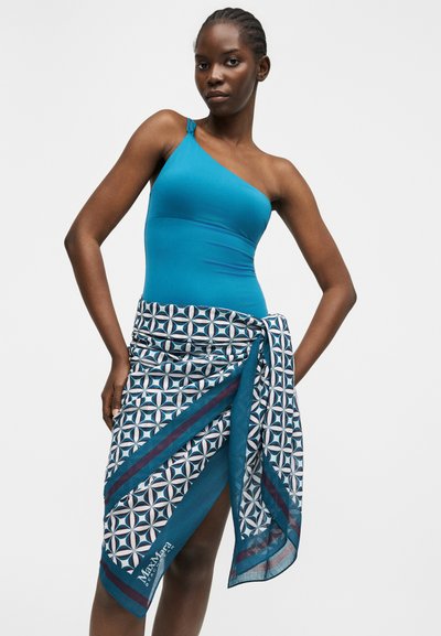 Γυναίκα με μοτίβο στην παραλία, φοράει πράσινο ολόσωμο μαγιό με έναν ώμο και ένα παρεό δεμένο στη μέση, στεκόμενη με τα χέρια στη μέση πάνω σε απλό φόντο.