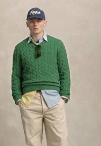 Groene kabelgebreide trui, lichtblauwe en gele overhemden eronder, khaki broek en een marineblauwe pet met het "Ralph" logo.