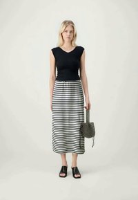 Femme portant un haut sans manches noir, une jupe midi rayée noir et blanc, des chaussures ouvertes noires, tenant un sac à main texturé noir à ses côtés.