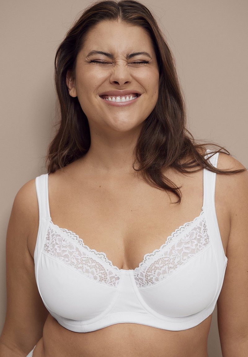 Vrouw met lang bruin haar, gekleed in een witte met kant afgewerkte bh, glimlachend met gesloten ogen voor een effen beige achtergrond.