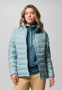 Junge Frau in einer hellblauen gesteppte Jacke über einem dunkelblauen Reißverschluss-Fleece, in beigen Hosen, vor einem einfarbigen grauen Hintergrund stehend.