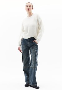 Weißer, gestrickter Pullover mit rundem Halsausschnitt, kombiniert mit blauen Denim- Cargo-Hosen mit Seitentaschen und lockerer Passform. Schwarze Schuhe.