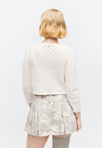 Femme aux cheveux blonds courts portant un haut blanc en maille à manches longues et une mini-jupe plissée grise à fleurs, vue de dos.