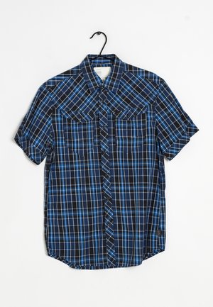 Chemise à manches courtes à carreaux bleue et noire avec deux poches poitrine, suspendue à un cintre noir sur un fond blanc.