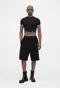Homme aux cheveux blonds courts portant un t-shirt court noir avec le texte « Masallah », un short large noir et des bottes noires mi-mollet, debout devant un fond blanc.