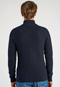 Marinblå stickad tröja med diamantmönster, ribbad krage och ärmslut, sedd bakifrån med långa ärmar och åtsittande design.