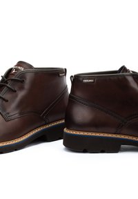 Botas de cuero marrón hasta el tobillo con punta redonda, cordones en la parte delantera y acentos azules contrastantes a lo largo de la suela. Detalles de costura y logotipo de la marca visibles.