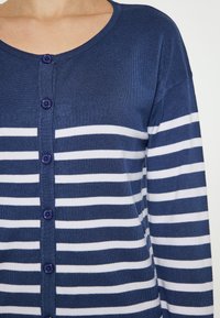 Mörkblå cardigan med vita horisontella ränder, ribbad textur, rund halslinning och lila knappar som löper längs fram.