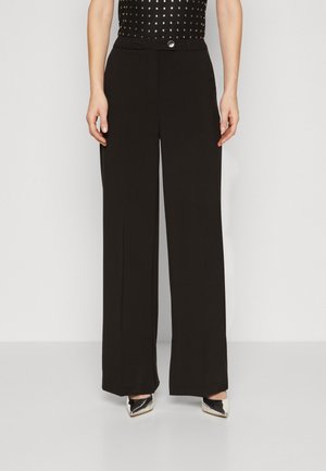 Woman wearing black wide-leg trousers with a buttoned waistband, paired with silver pointed-toe heels and a studded black top.