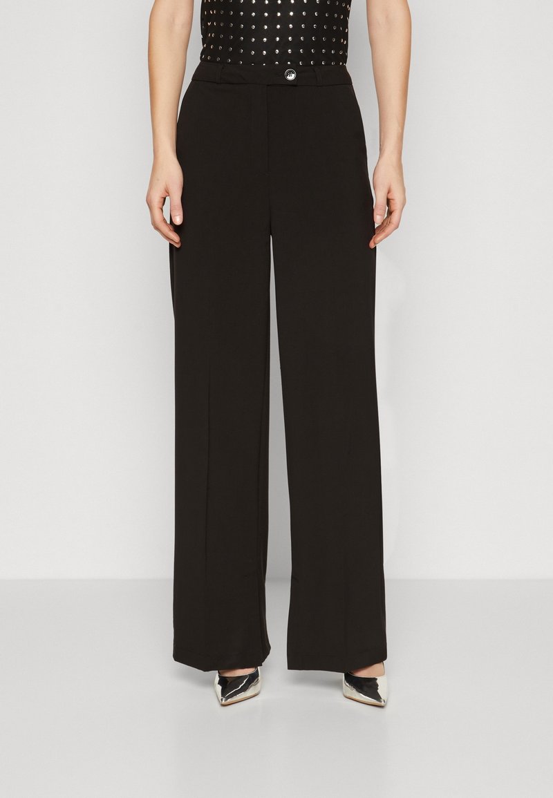 Femme portant un pantalon noir à jambes larges avec une ceinture boutonnée, associé à des escarpins argentés à bouts pointus et un haut noir clouté.