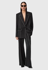 Vrouw in een zwart op maat gemaakt pak met witte stiksels, oversized blazer, wijde broek, zwarte puntige schoenen en een zonnebril, staand met één hand in de zak.
