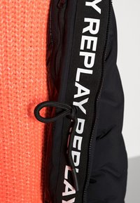 Detailní záběr na černou bundu s bílým logem REPLAY v oblasti zipu vedle strukturované oranžové pletené látky.