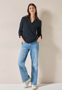Blusa navy testurizzata con scollatura a V, abbinata a jeans larghi di colore azzurro chiaro. Sneakers bianche completano il look casual. Sfondo semplice.