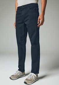 Marineblauwe denim jeans met een rechte pasvorm, uitgerust met een knoopsluiting, riemlussen en vijf zakken. Draag met grijze sneakers.
