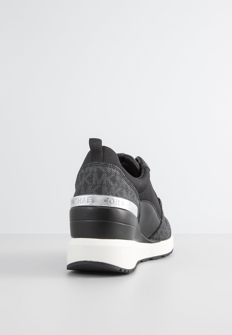 Zwarte sneaker met textuurstof, elastische banden, een zilveren Michael Kors-embleem en een contrasterende witte tussenzool, met een slank ontwerp.