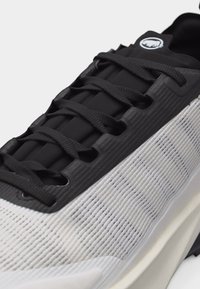 Κοντινό πλάνο μαύρου και λευκού αθλητικού παπουτσιού με υφασμάτινο πάνω μέρος με υφή, μαύρα κορδόνια και κυκλικό λογότυπο στη γλώσσα.