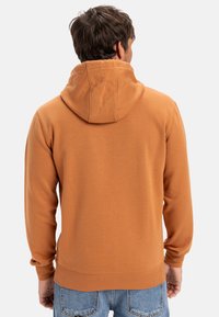 Orange bomuldshoodie med snoretræk i hætten, ribbede manchetter og kant, vist bagfra, parret med lyseblå jeans.