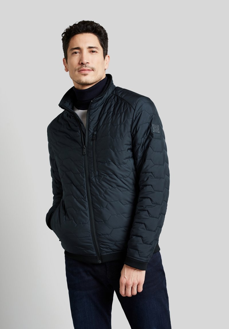 Veste matelassée en bleu marine foncé avec une fermeture éclair à l'avant, un col montant et un motif de couture hexagonal. Comprend des poches latérales et des poignets côtelés.