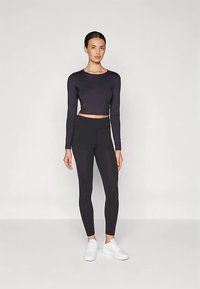 Egy nő áll, fekete hosszú ujjú crop toppot, fekete leggingset és fehér sportcipőt visel, egy világos, egyszínű háttér előtt.