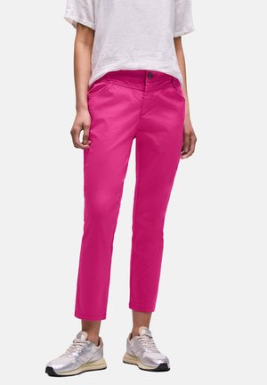 Femme portant un pantalon slim rose vif, des baskets blanches décontractées et un haut blanc ample à manches courtes, debout sur un fond uni.