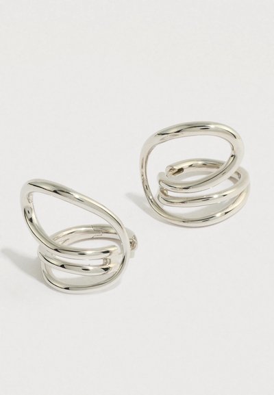 Ζευγάρι από γυαλιστερά σκουλαρίκια κρίκους από ασημένιο μέταλλο με γλυπτικό, ανοιχτό, αλληλοεπικαλυπτόμενο, ακανόνιστο οβάλ σχέδιο σε λευκό φόντο.