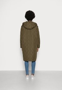 Cappotto trapuntato verde oliva con cappuccio e silhouette lunga e dritta. Presenta motivi a diamante in rilievo e tasche laterali cucite, abbinato a jeans blu e scarpe da ginnastica bianche.