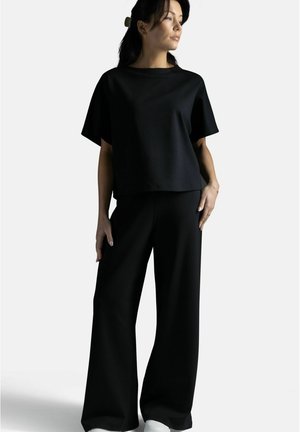 Nainen poseeraa löysässä mustassa lyhythihaisessa topissa ja leveälahkeisissa mustissa housuissa, katsomassa hieman sivulle yksinkertaisen vaalean taustan edessä.