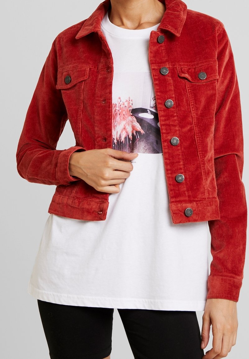 Rote Cordjacke mit taillierter Passform, ausgestattet mit schwarzen Knöpfen und Brusttaschen, kombiniert mit einem weißen T-Shirt mit Grafikmotiv darunter.