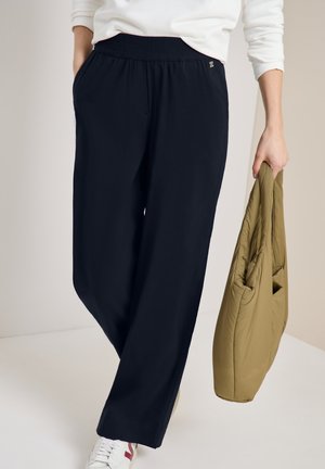 Personne portant un pantalon noir à taille élastique, des baskets blanches et un haut blanc, tenant un sac fourre-tout matelassé vert olive.