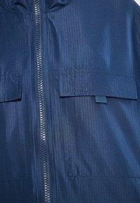 Temno modra jakna iz gladkega, teksturiranega materiala ima sprednje zapiranje na zadrgo, dva žepa na prsih in kapuco. Vidna je navpična rebrasta struktura.