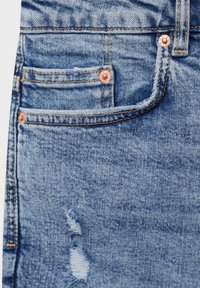 Tissu en denim bleu clair avec un design vieilli. Présente des rivets de couleur cuivre, un style cinq poches et des détails de couture visibles.