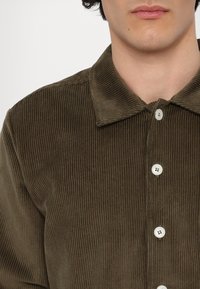 Rjava kordurojska srajca z klasičnim ovratnikom, horizontalno rebrasto teksturo in belimi gumbi po sprednji strani.
