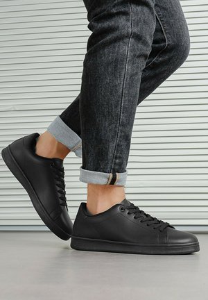 Personne portant un jean en denim foncé retroussé et des baskets noires basses, debout sur un sol gris avec un mur à rayures en arrière-plan.