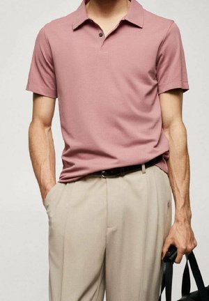 Mand iført støvet lyserød kortærmet poloshirt, stoppet ned i beige bukser med sort bælte, holder en sort taske i højre hånd.