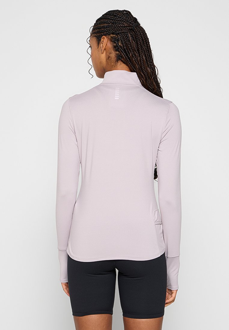 Femme aux cheveux tressés portant un haut à manches longues violet clair et un short de cyclisme noir, debout de dos face au spectateur.