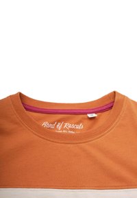 Luonnollisesta puuvillasta valmistettu t-paita, jossa on pyöreä kaula-aukko. T-paidassa on poltettu oranssi väri, valkoinen raita ja vaaleanpunainen sisäkaulus. "Band of Rascals" -logo näkyy.