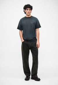 Zwart T-shirt met korte mouwen en een klein grafisch zakje, gecombineerd met wijde donkergroene broek en zwarte schoenen op een effen achtergrond.