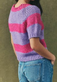 Persona con un suéter de punto grueso de manga corta con rayas horizontales rosas y moradas, combinado con jeans de denim azul.