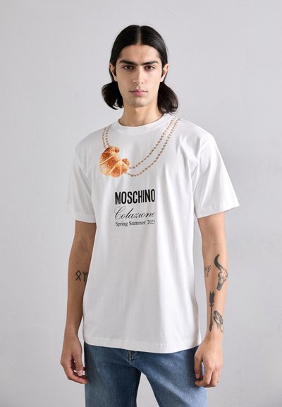 T-shirt bianca in cotone con grafica di un croissant e design di una collana di perle, con testo "Moschino Colazione Primavera Estate 2023" sottostante.