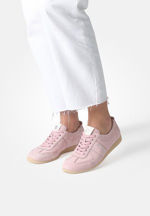 Feet wearing light pink lace-up sneakers with beige soles, paired with cropped white pants with frayed hems.